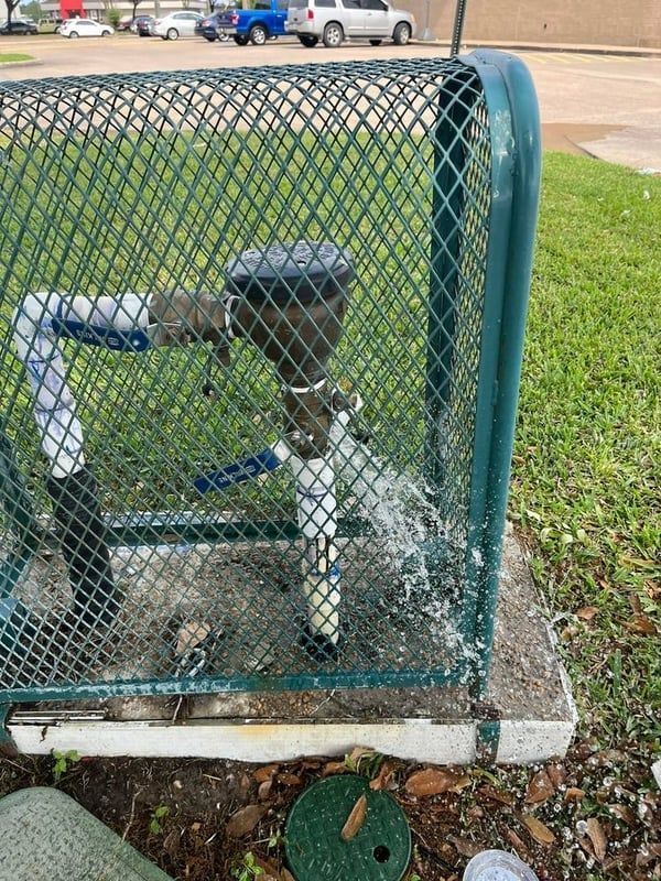 Water meter assembly behind a green chain-link fence on a concrete pad near a grassy roadside.