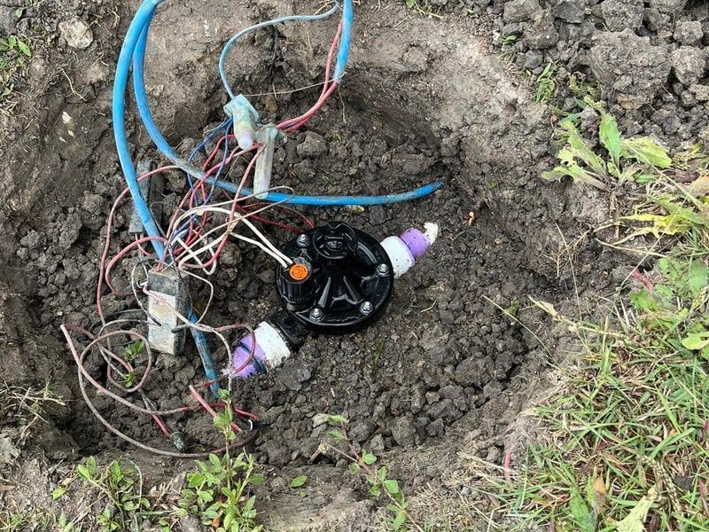 Exposed wires and a black sprinkler valve buried in a muddy ground hole.