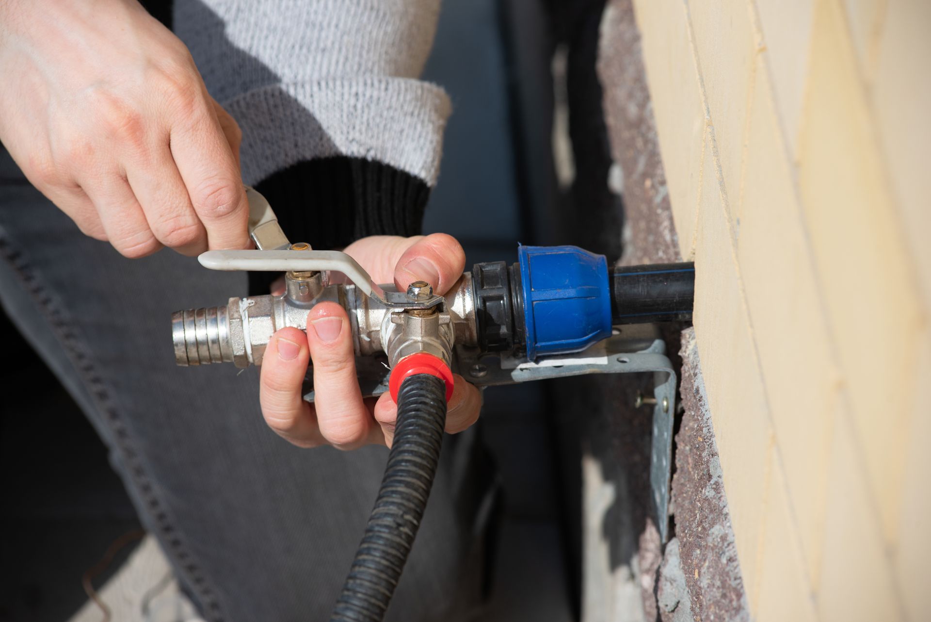 Hands adjusting a metal pipe fitting with a wrench near a wall, with a blue connector and red hose attached