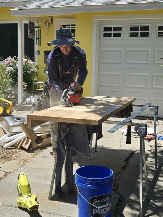 Man in overalls and hat using a circular saw on a wood board outdoors. Yellow house in background.