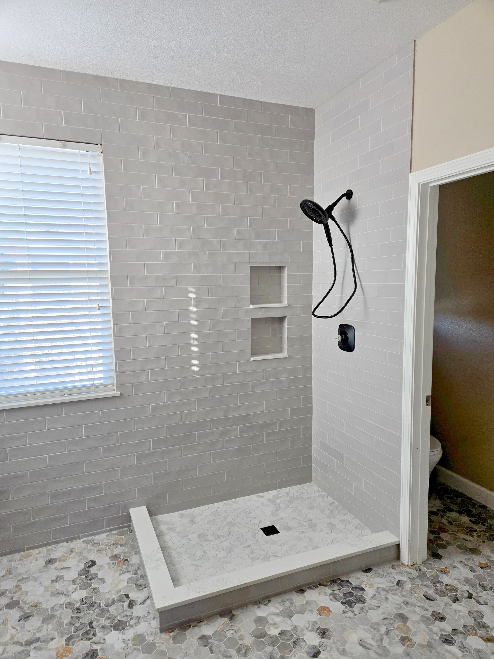 Bathroom with gray tile shower and floor, shower head, window with blinds, and open doorway.