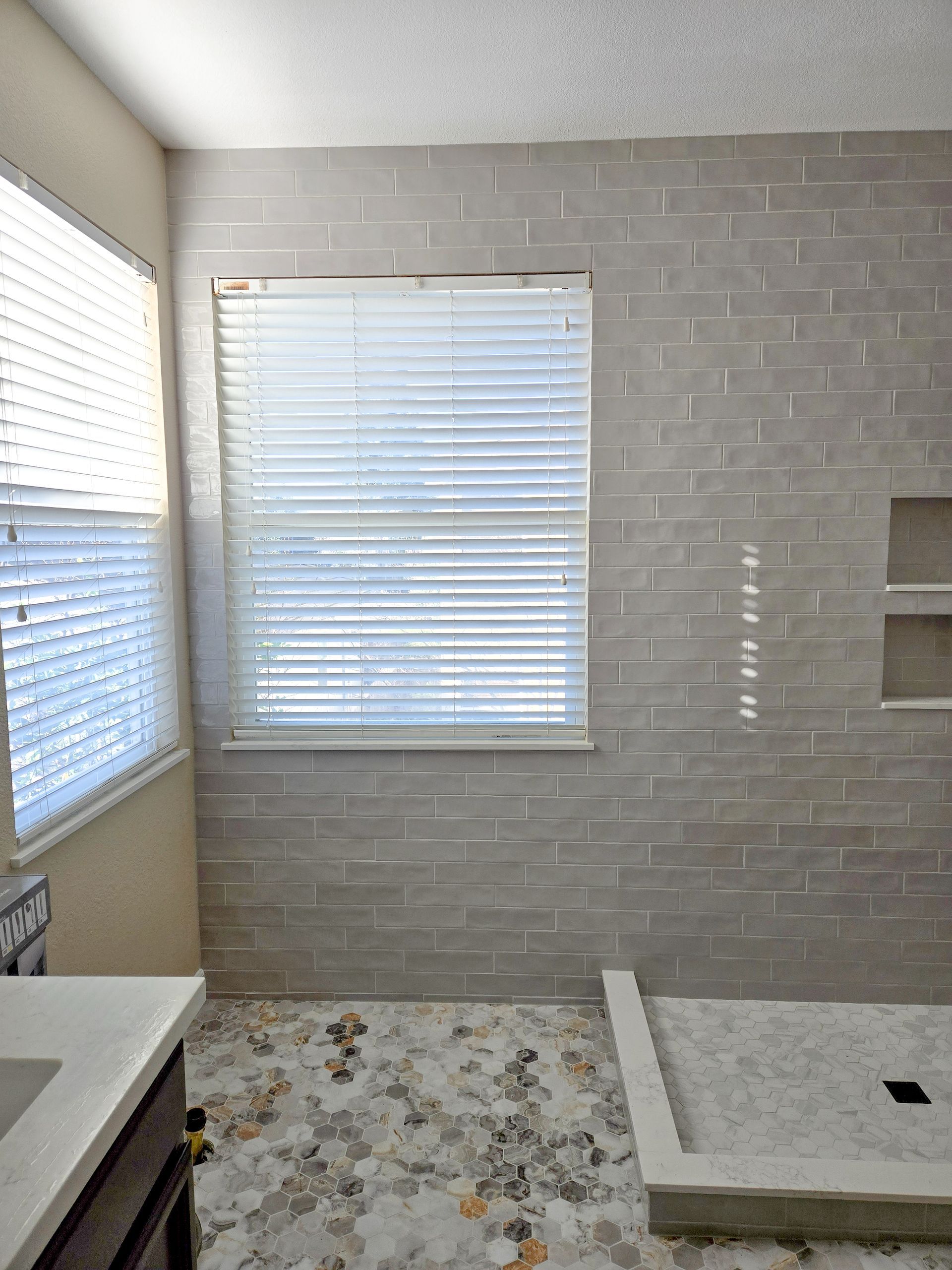 Bathroom with gray brick-pattern tile, a window with blinds, and a shower area.