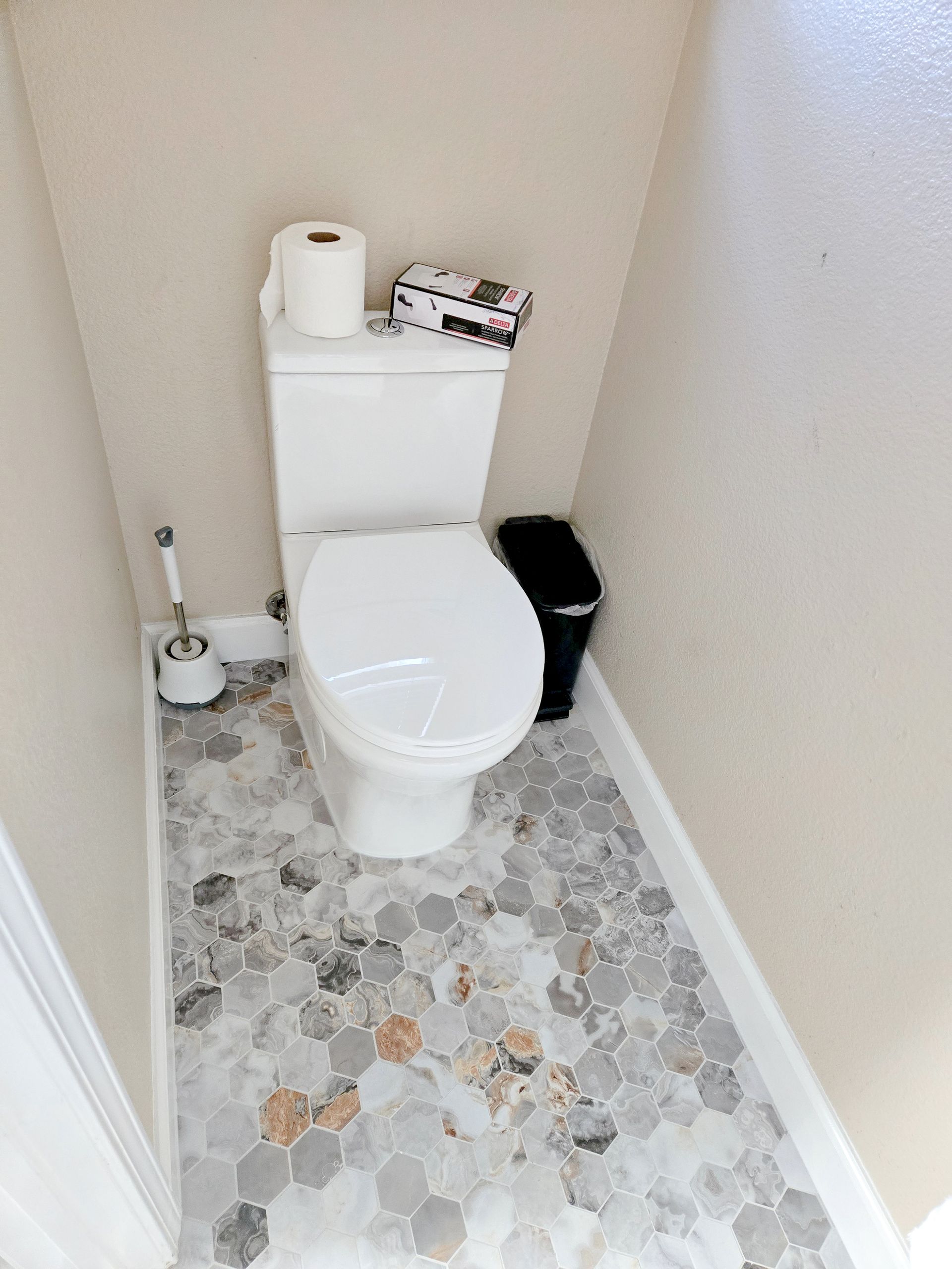 Toilet in a small bathroom with patterned tile floor, toilet paper, and a trash can.