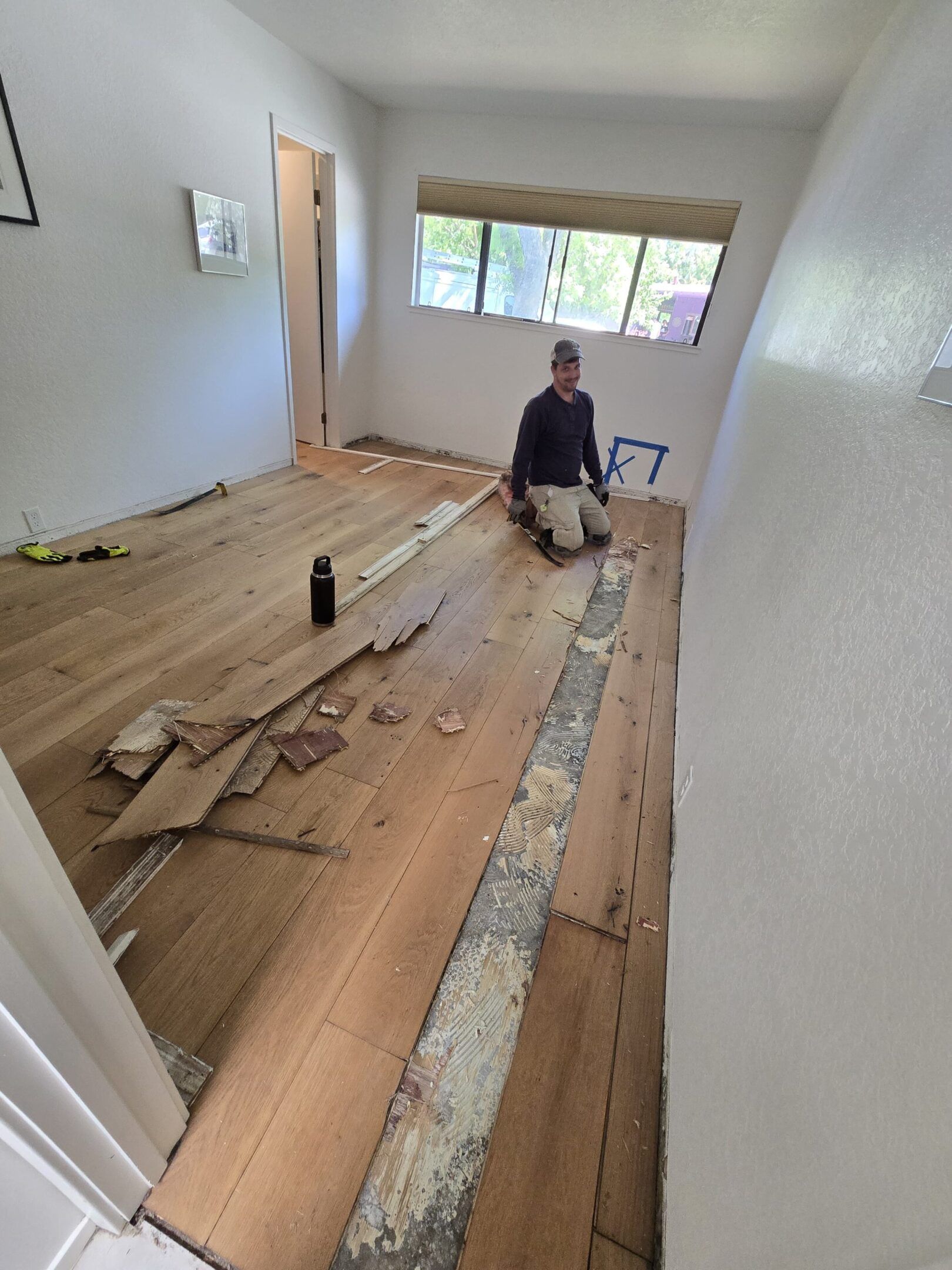 Person removing flooring in a room with white walls, a window, and an open doorway.