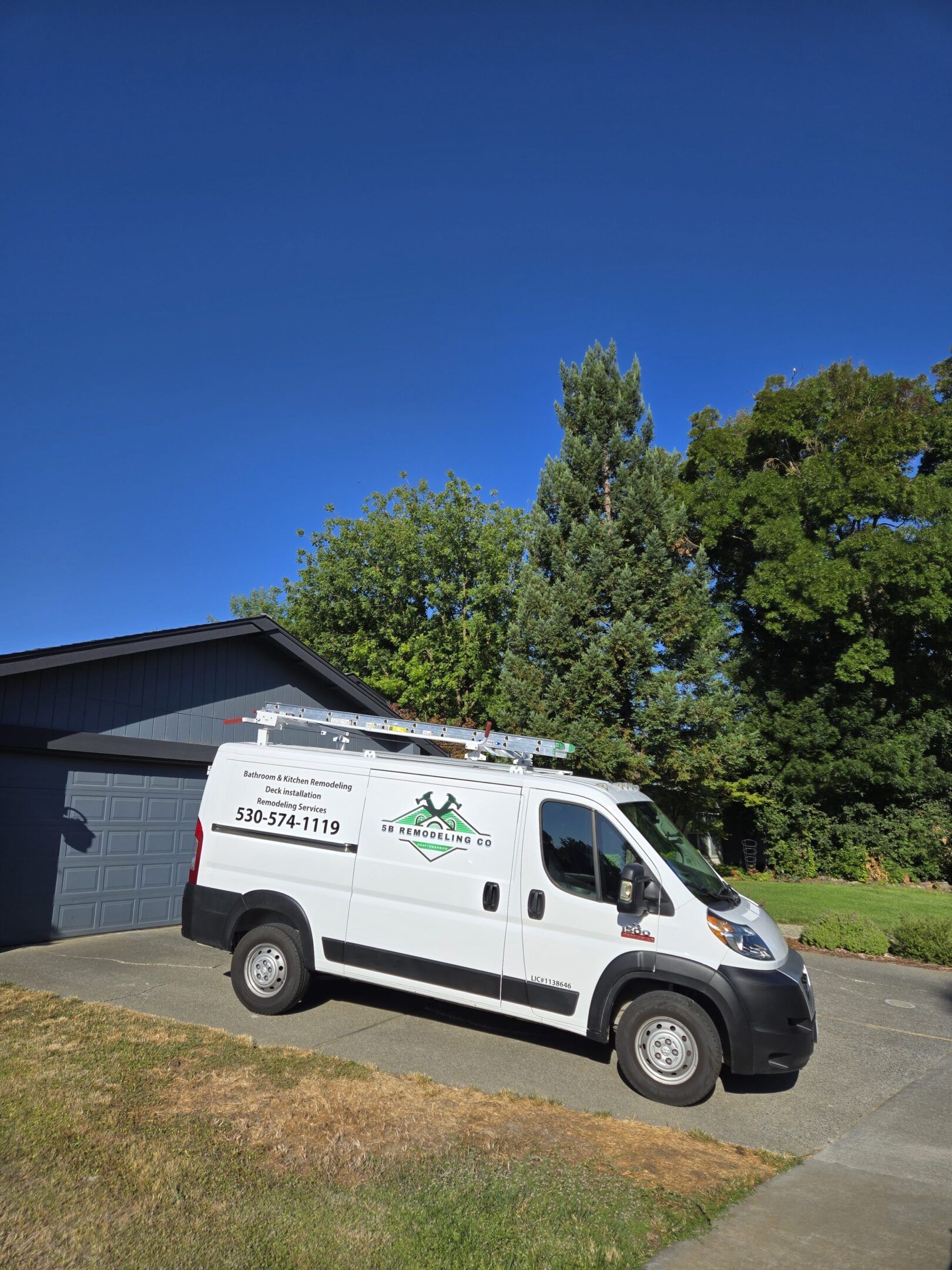 White van parked in a driveway in front of a blue-gray house. Green trees and a clear blue sky.