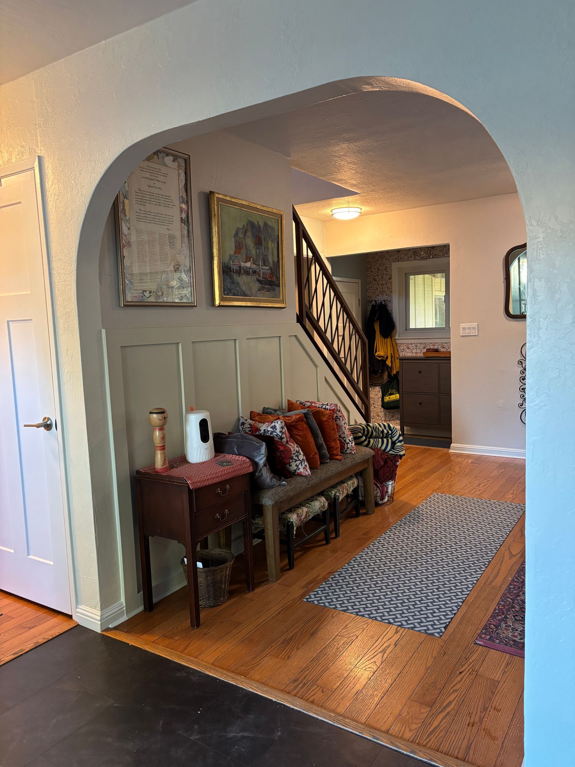 Archway leading into a home with a bench, artwork, and staircase. Cork flooring and gray and white walls.