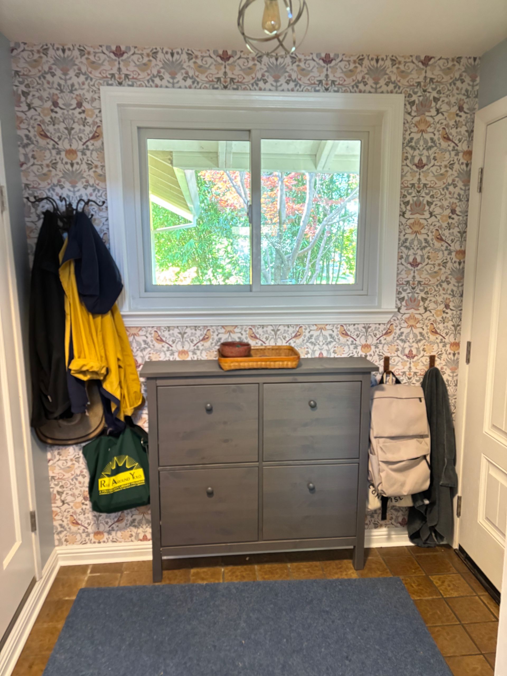 Entryway with gray shoe cabinet, patterned wallpaper, window, and coats hanging on hooks.