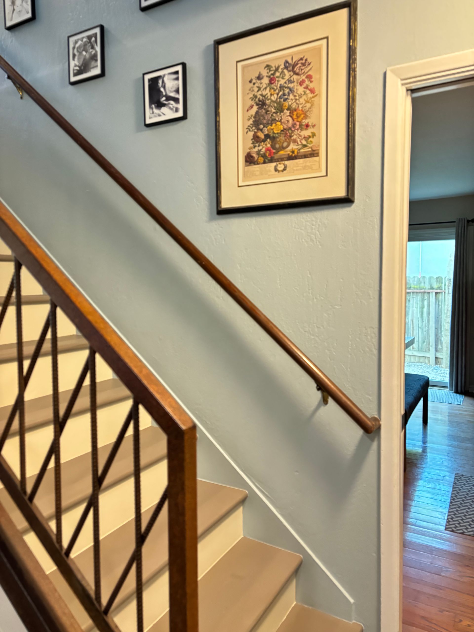 Staircase with wooden handrail and banister. Light blue wall with framed artwork. Doorway to another room.