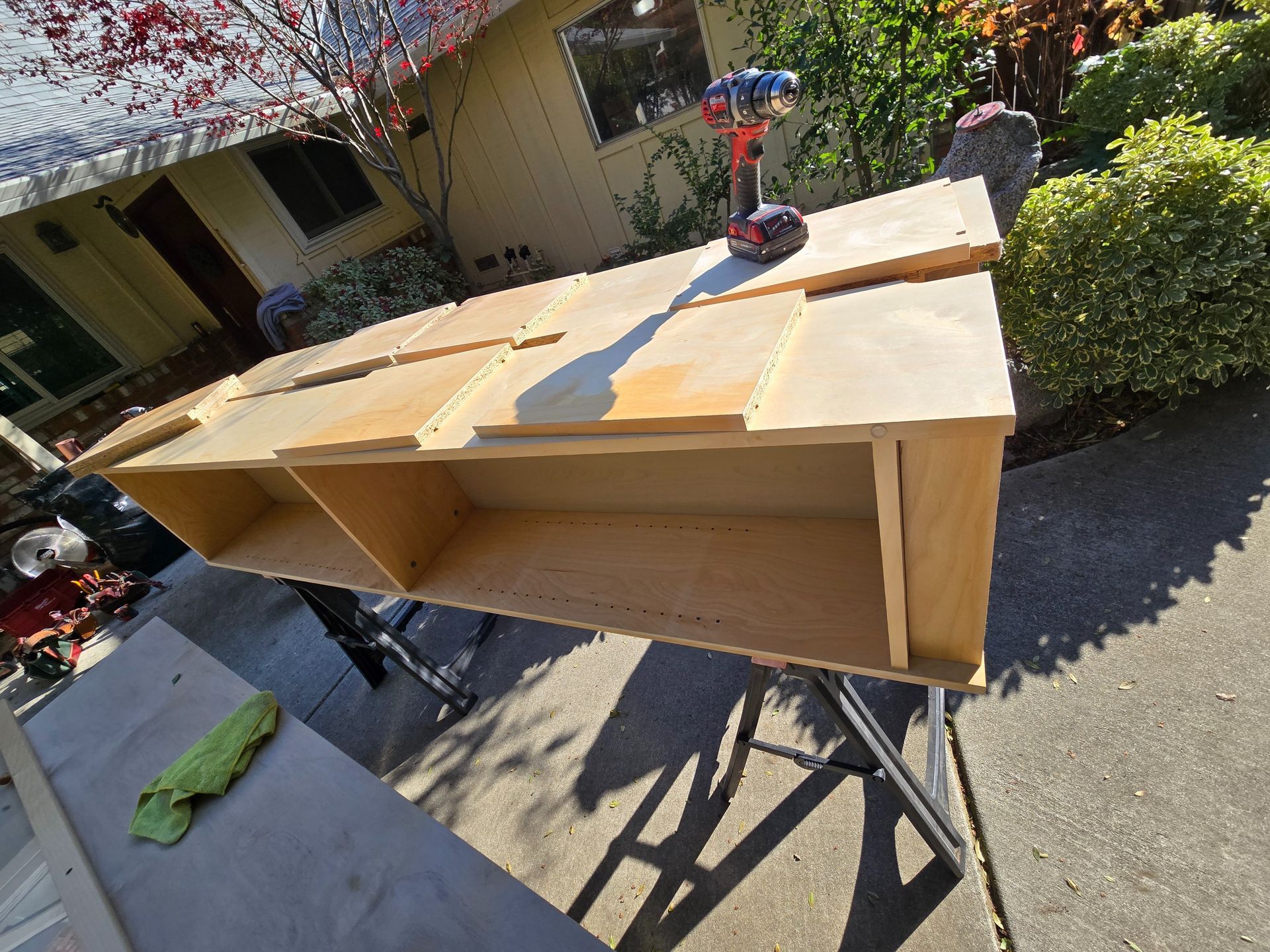 A partially constructed wooden cabinet on sawhorses outdoors with a drill on top.