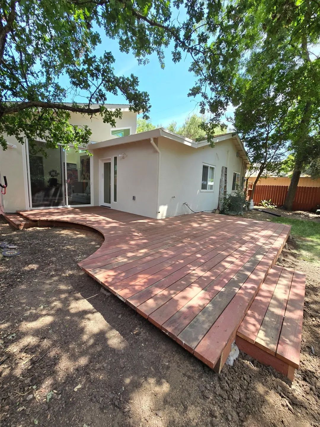Backyard deck with reddish-brown wood, steps, and house with glass doors. Trees overhead and dirt ground.