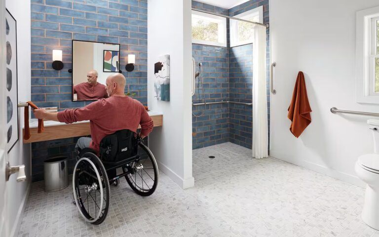 Man in wheelchair in accessible bathroom, reaching for towel; blue tile, grab bars, open shower.