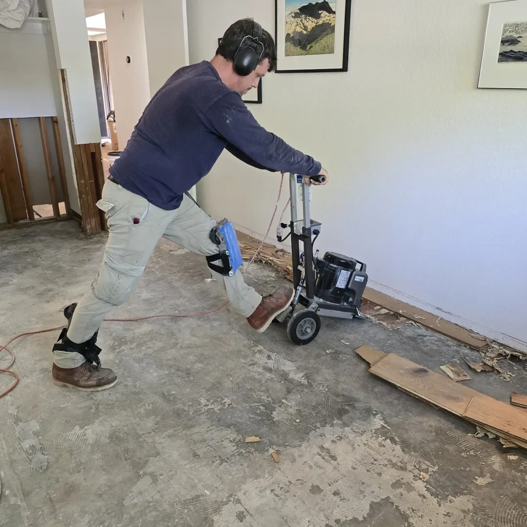 Man sanding a concrete floor with a floor sander, wearing knee pads and ear protection.