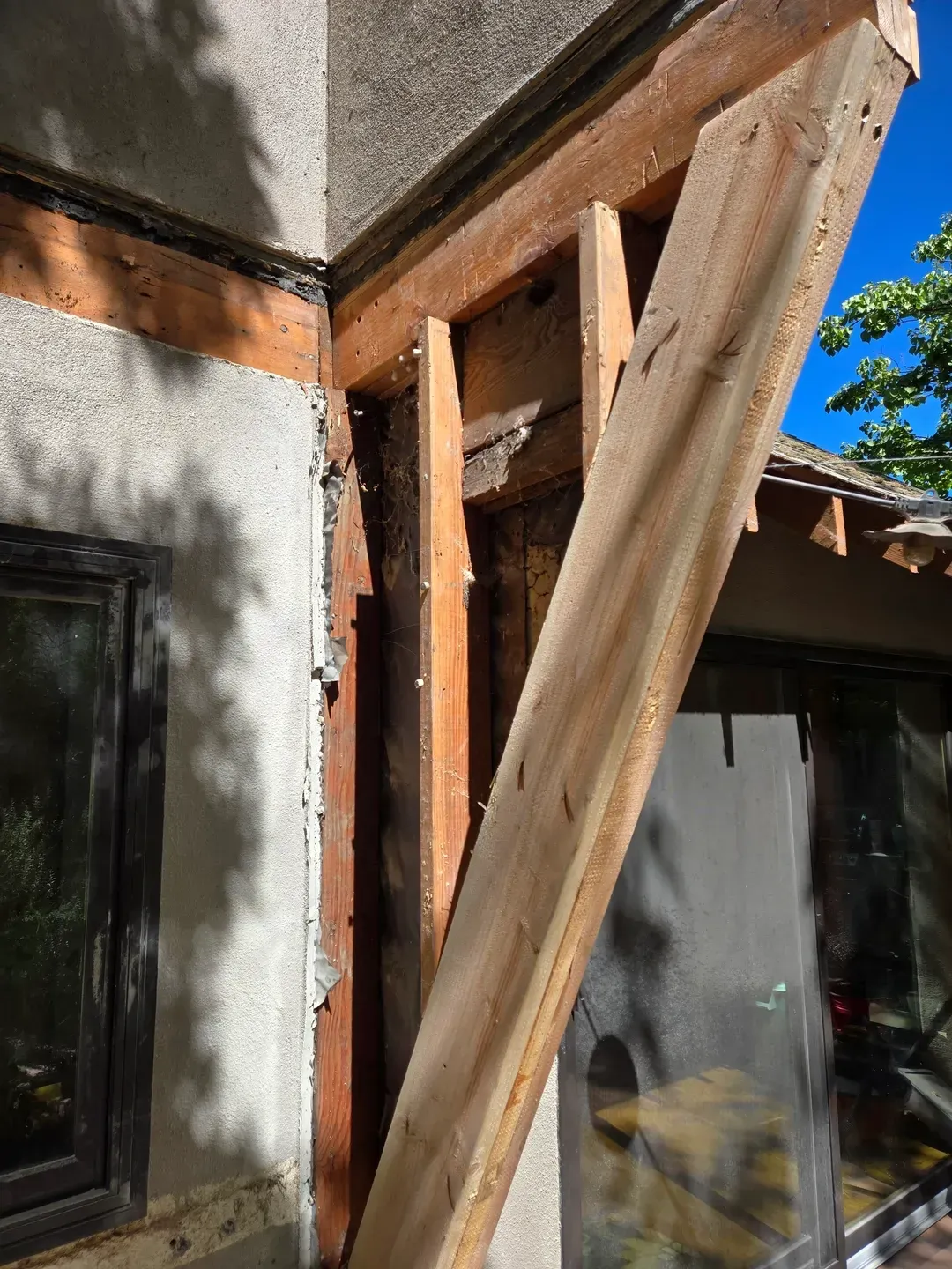 Wood beam bracing corner of a building with stucco and a window. Brown wood, gray stucco, sunny day.