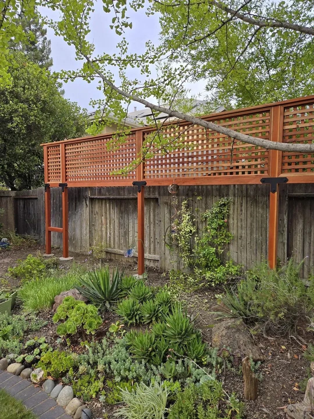 Wooden deck with latticed railing over a garden and a wooden fence. Brown and green colors.