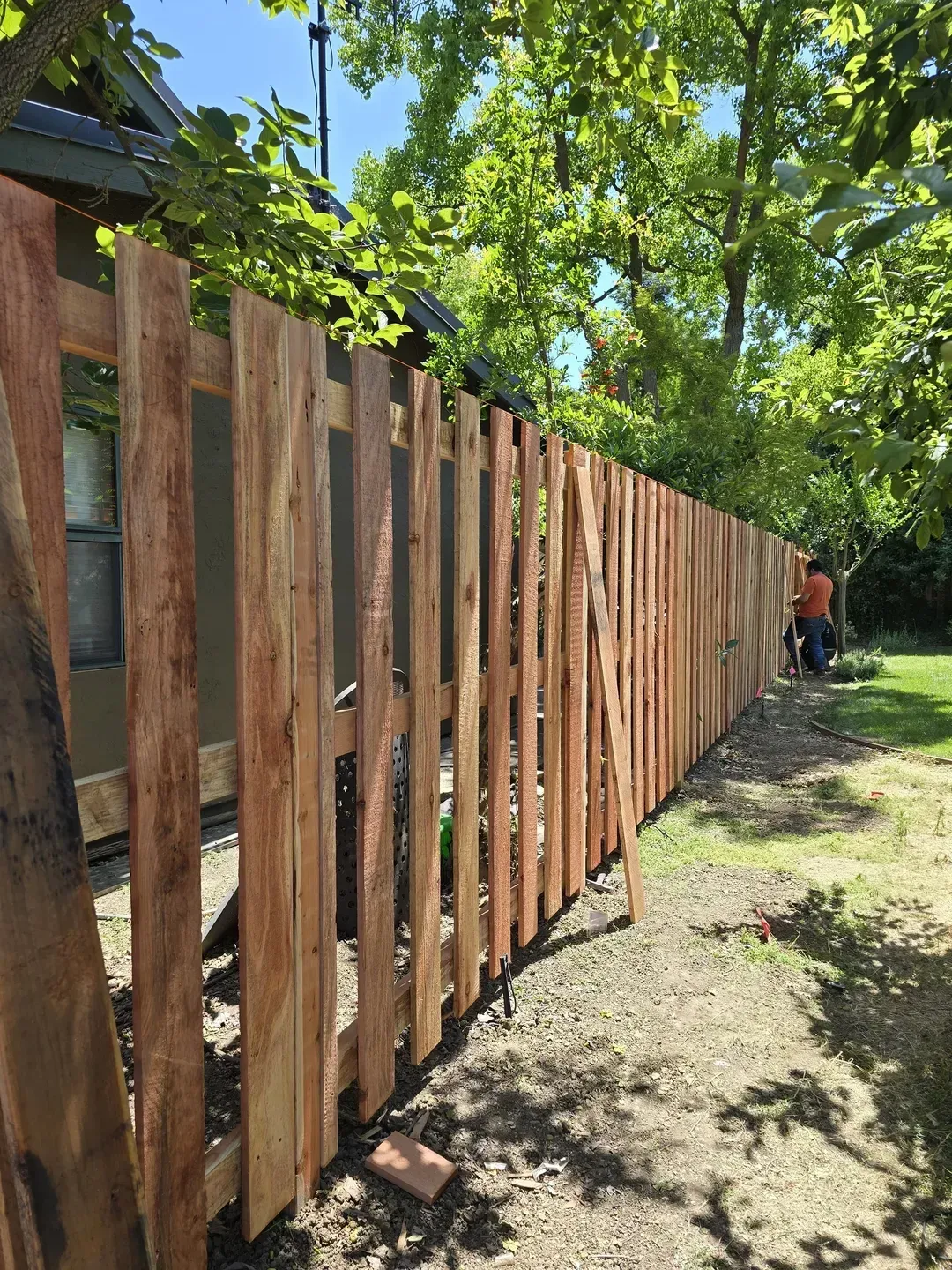 A newly built wooden fence in a yard with a person walking in the distance, surrounded by trees.