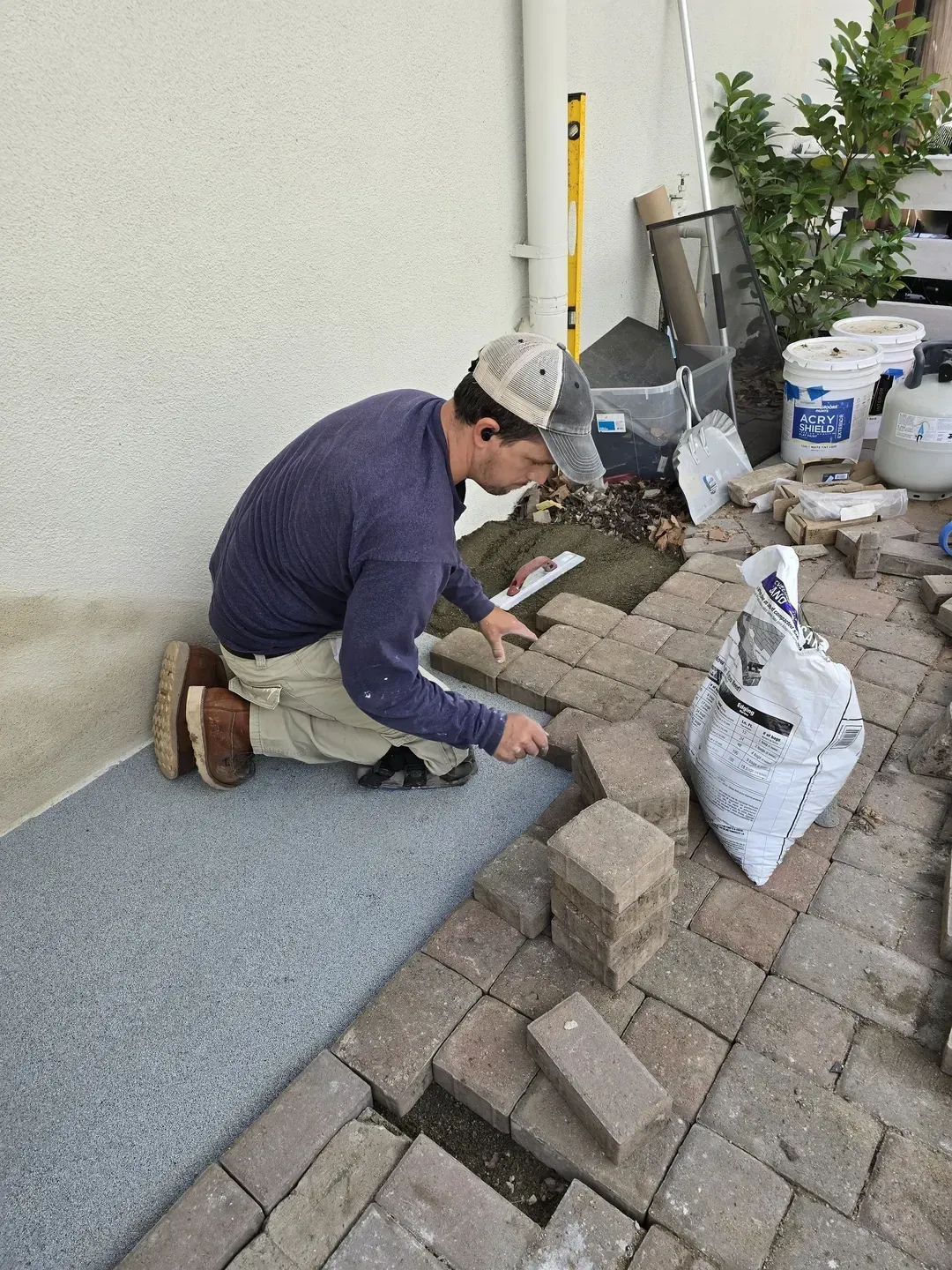 A person kneels, laying brick pavers next to a gray patio section near a white wall.