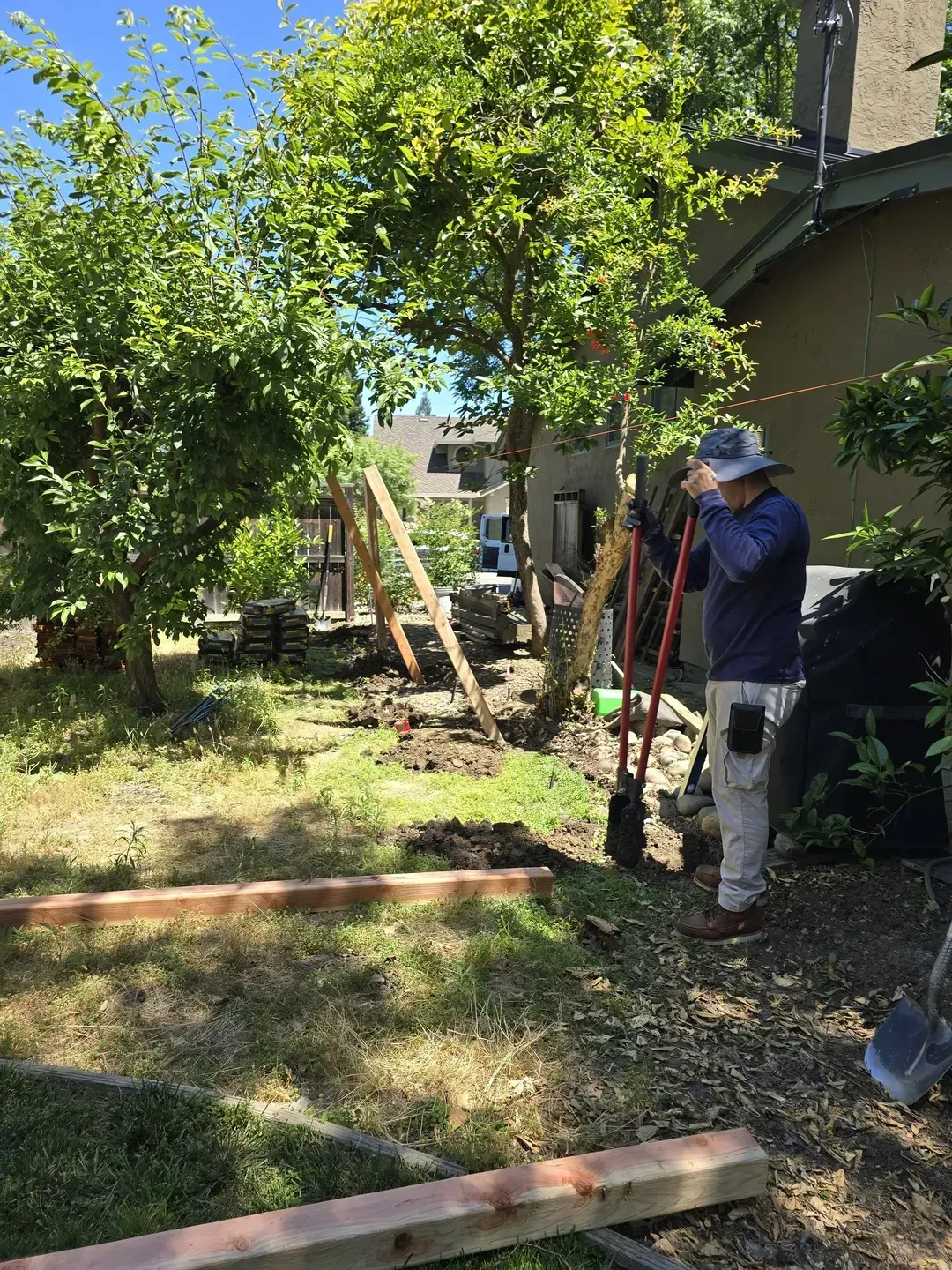 Person in hat uses tools to work on a fence in a backyard. Trees and building visible.