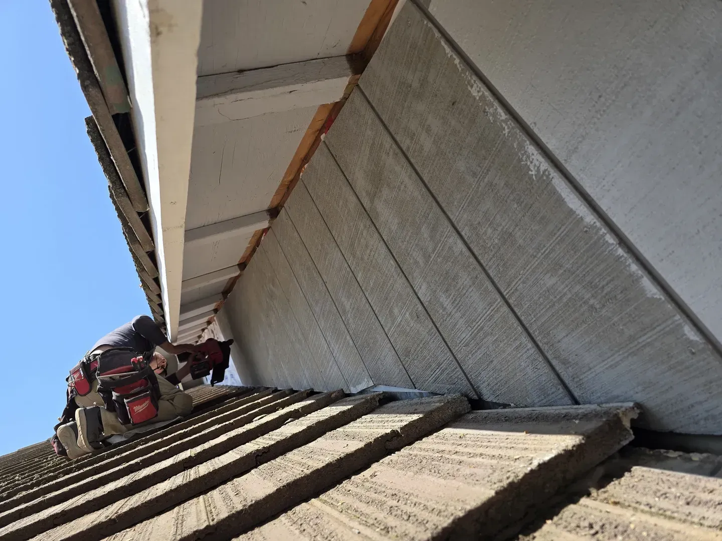 Person on a roof installing gray siding on the side of a building, under a white overhang.