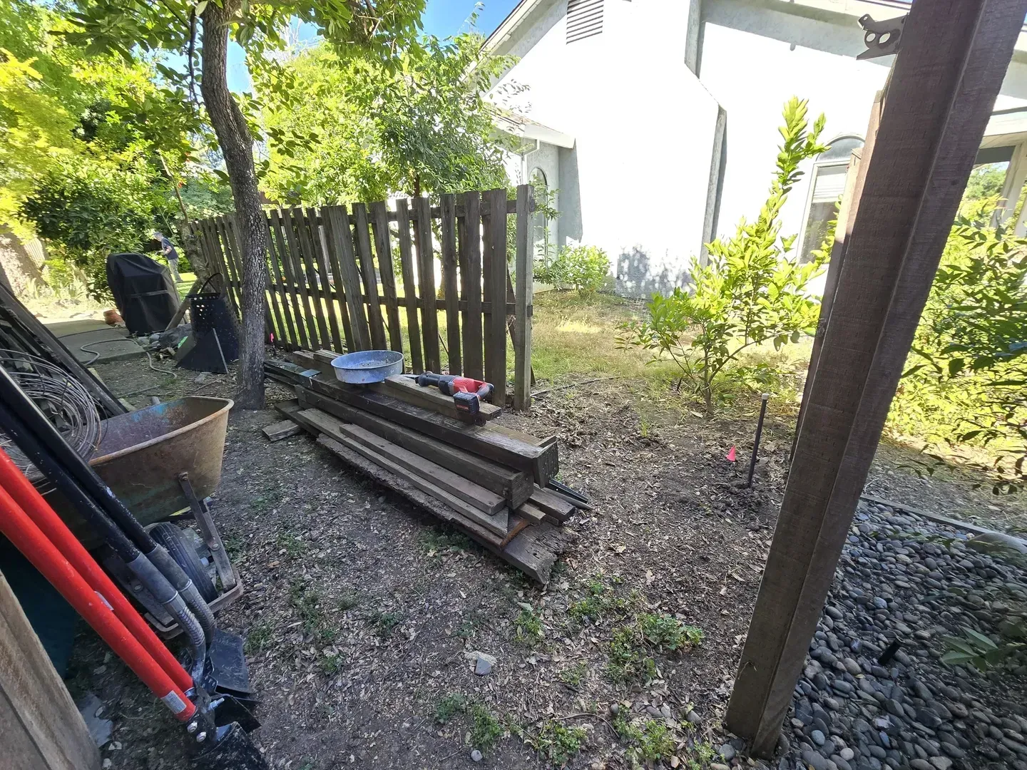 Backyard with wooden fence, tools, and lumber on gravel ground near a house.