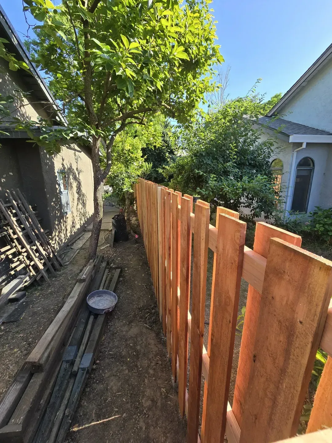 Newly built wooden fence along a narrow yard between two houses, with a tree in the background.