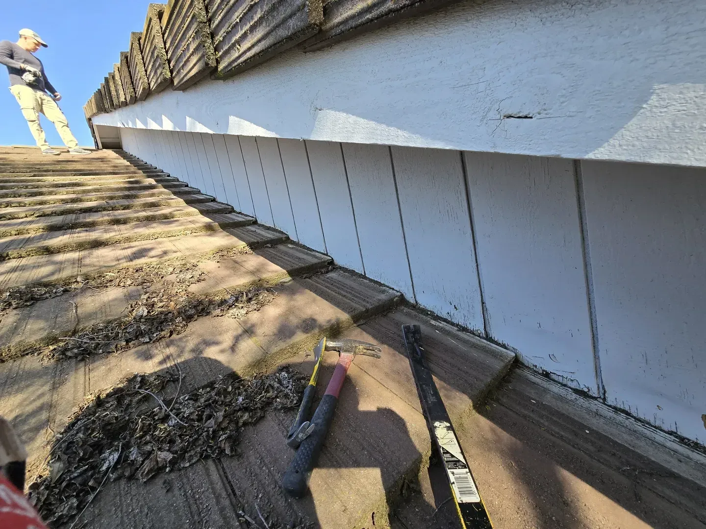 Person on roof repairing soffit; white paneling, brown roof tiles, sunny day.