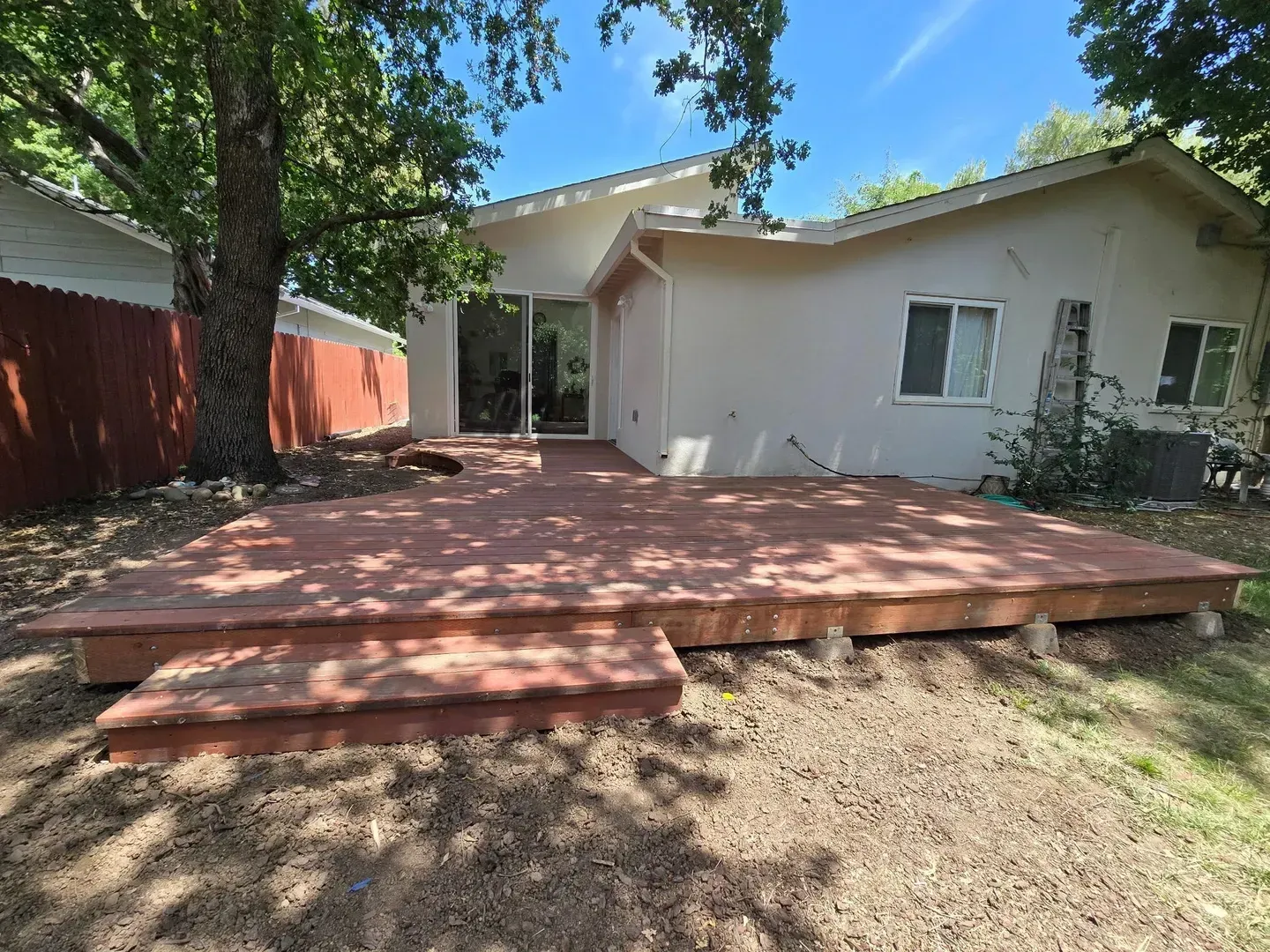 Backyard deck with steps, a house, and trees. Brown deck, red fence, gravel ground, and blue sky.