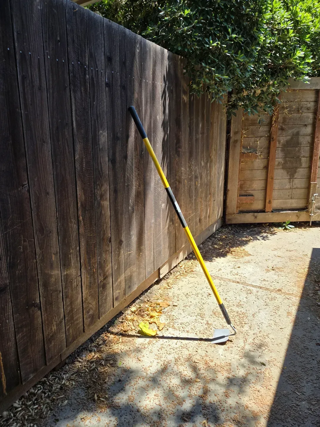 Yellow-handled garden hoe leans against a weathered wooden fence on a sunny patio, casting a shadow.