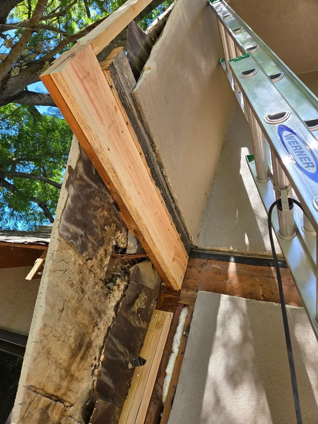Construction site, angled view: wooden beam attached to damaged stucco wall. A ladder leans against the building.