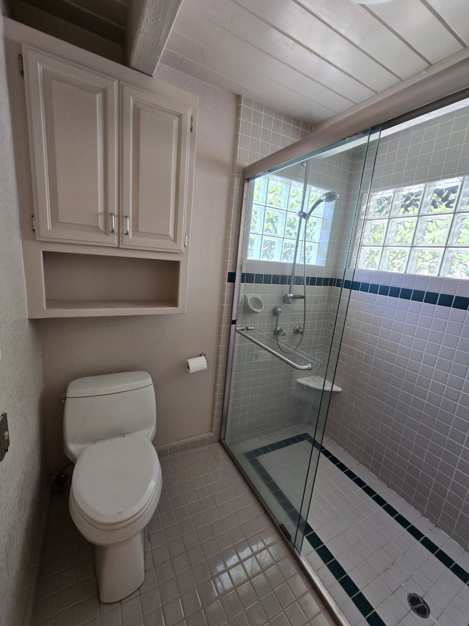 Bathroom with toilet, shower, and cabinets; light colors, small tile, glass shower door.