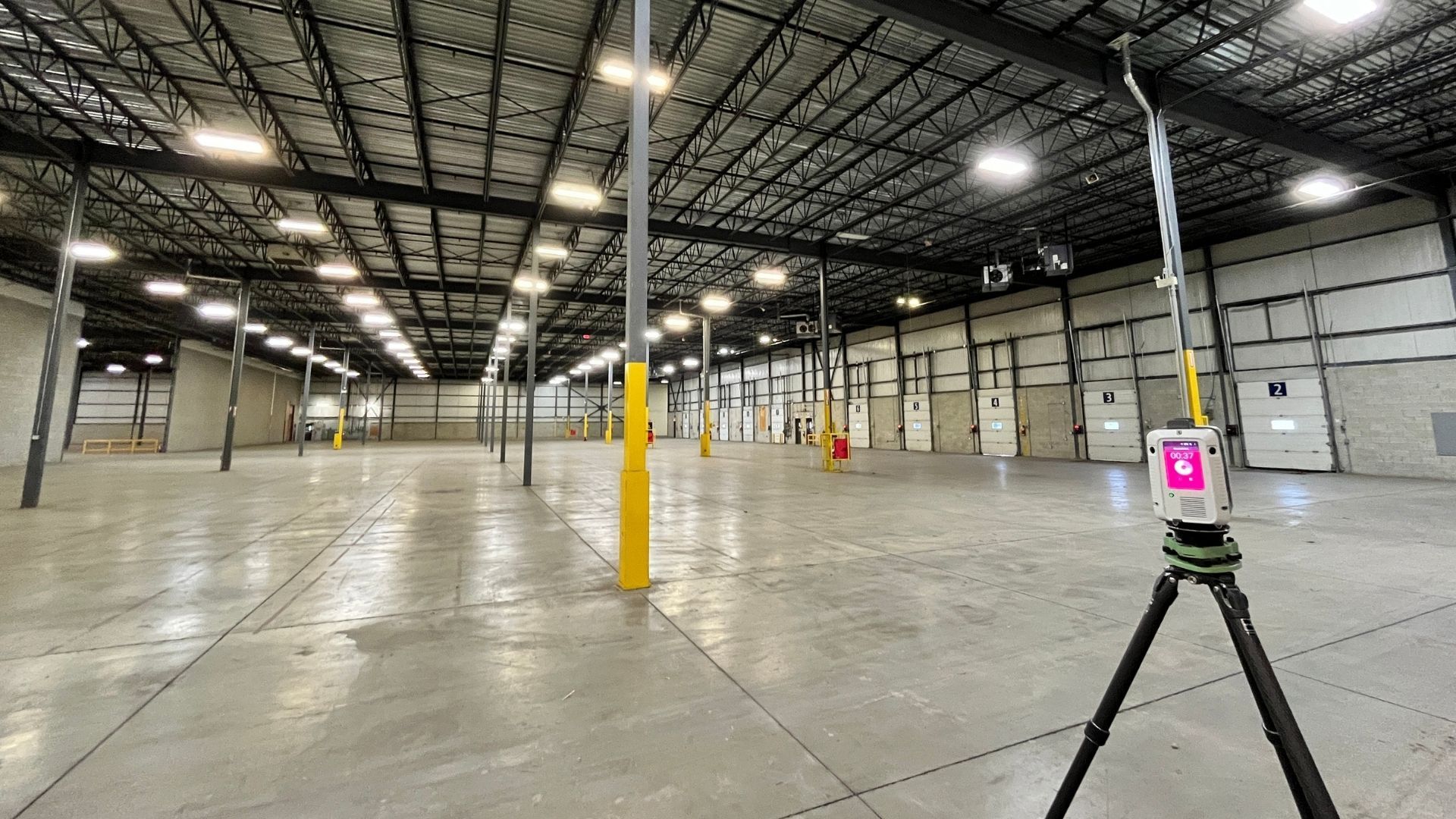 Tripod-mounted device in empty warehouse with high ceiling, gray floor, yellow support poles.