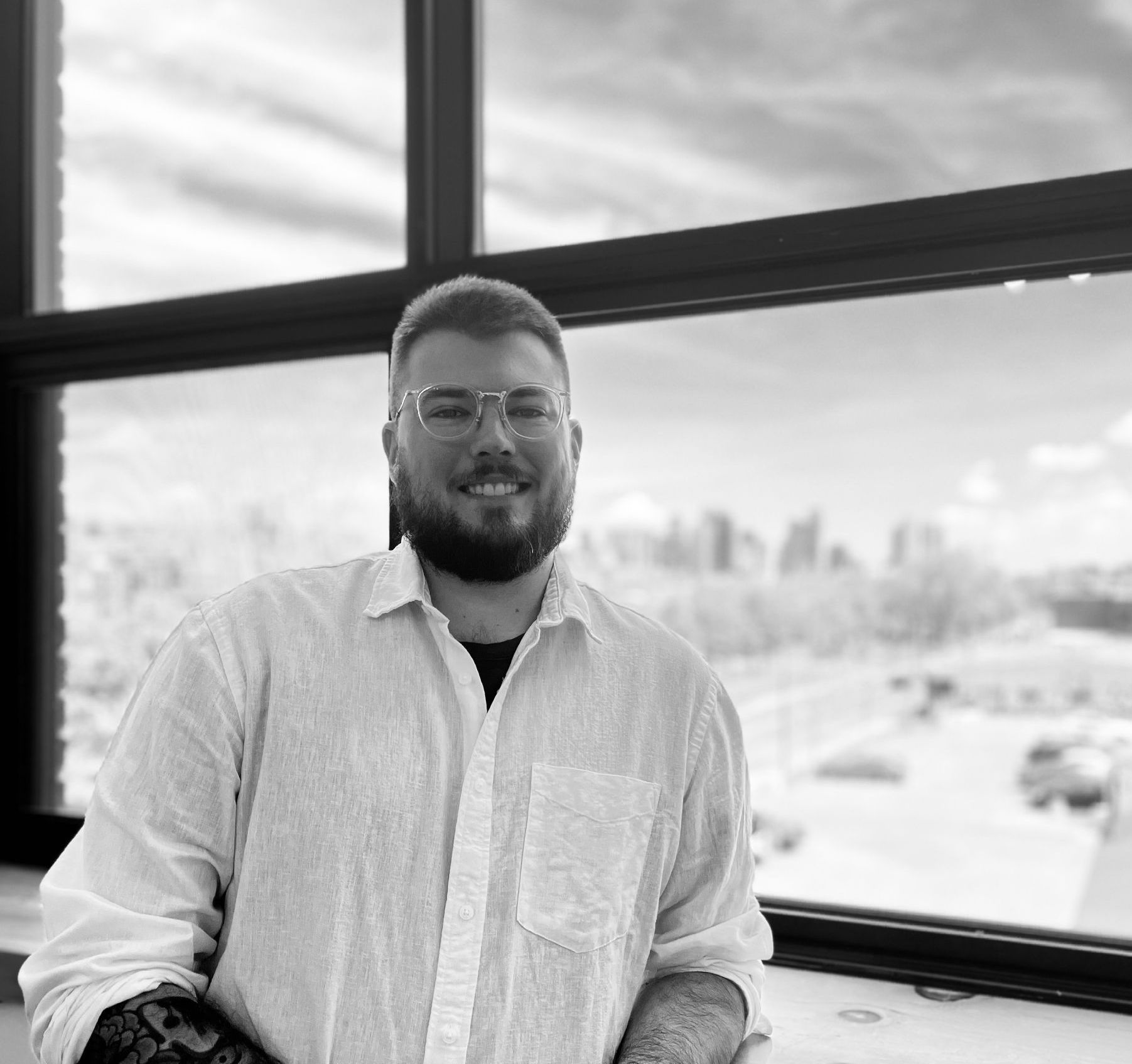 Man in glasses smiles, sits near a window with city view, wearing button-down shirt.