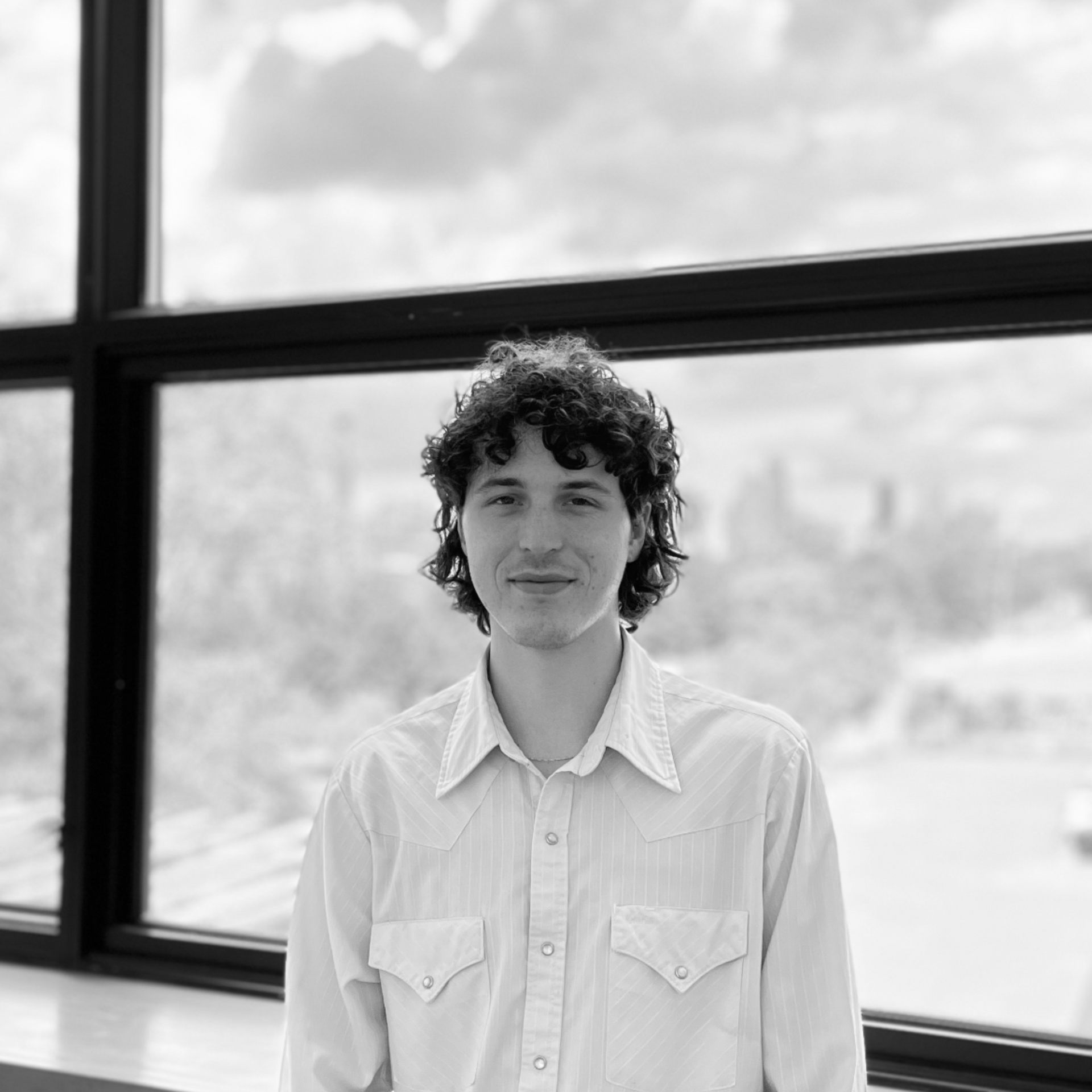 Man with curly hair in a light-colored button-down shirt stands in front of a window.