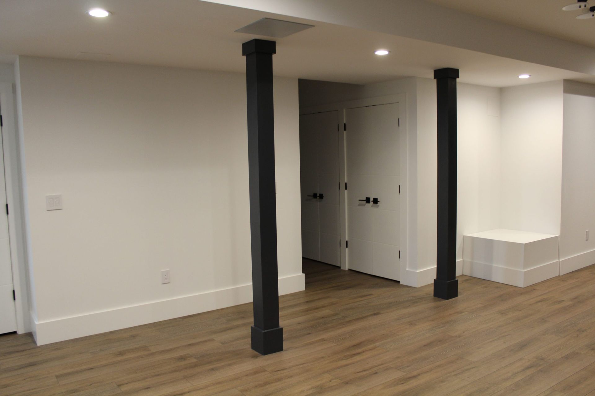 Minimalist basement hallway with white walls, dark beams, and wood flooring.