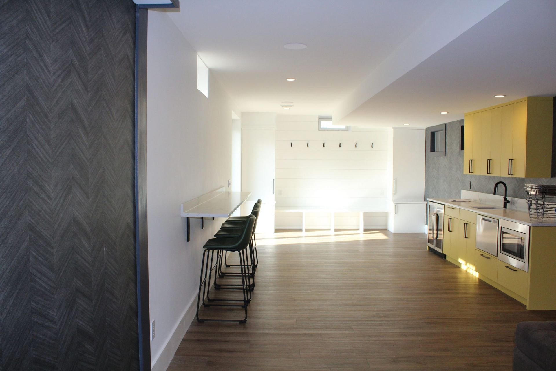 Basement kitchen with a long white bar, yellow cabinets, and hardwood floors. Green stools are lined up at the bar.