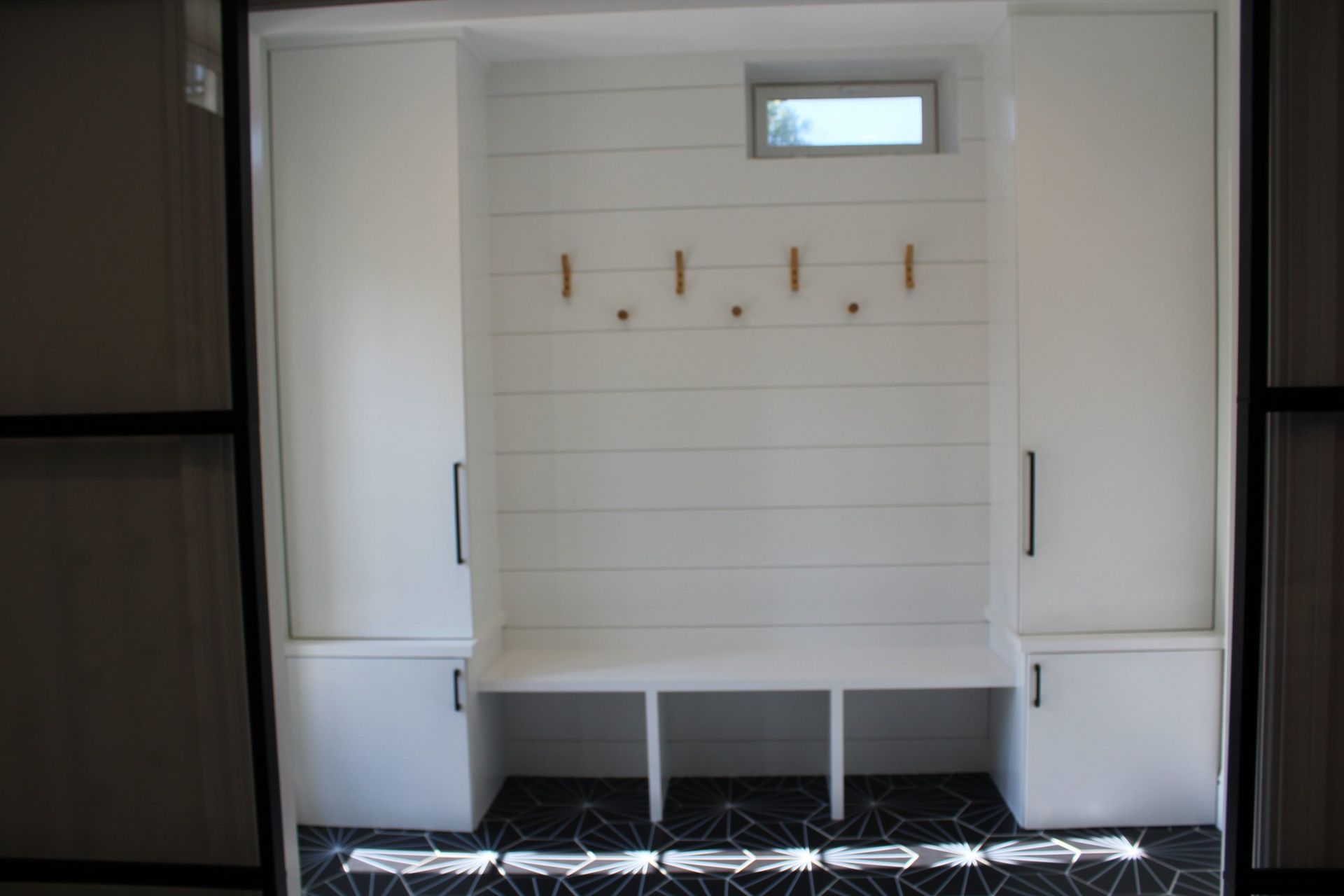 White entryway built-in with cabinets, bench, and hooks. Black and white patterned floor and small window.