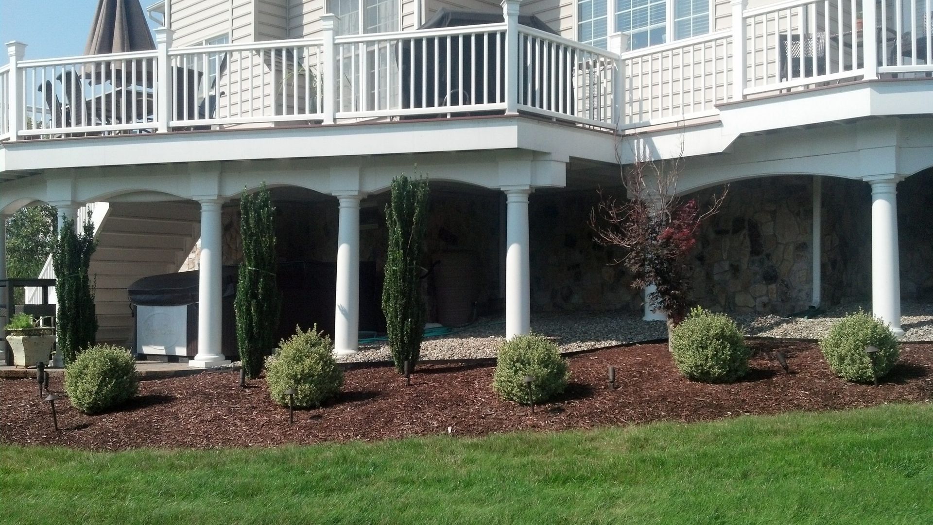 Under-deck area with white columns, stone wall backdrop, and landscaped shrubs.