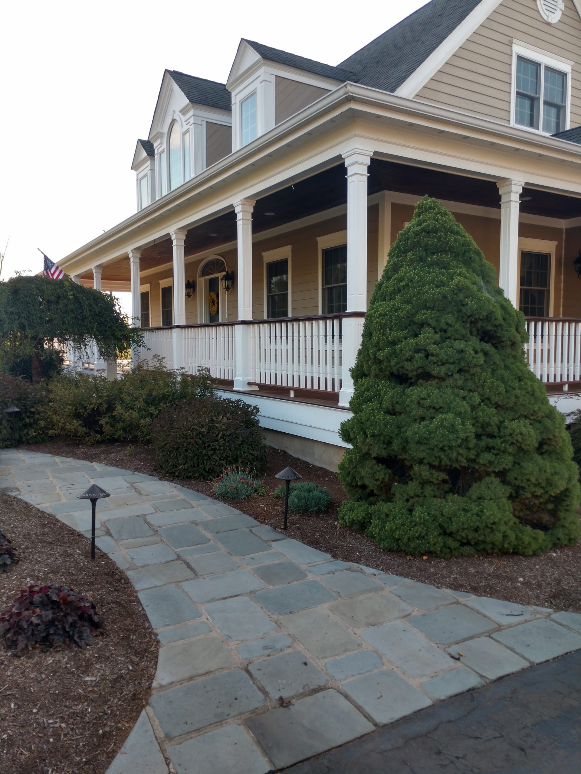 Front walkway to beige house with white trim, wraparound porch, and manicured shrubs.