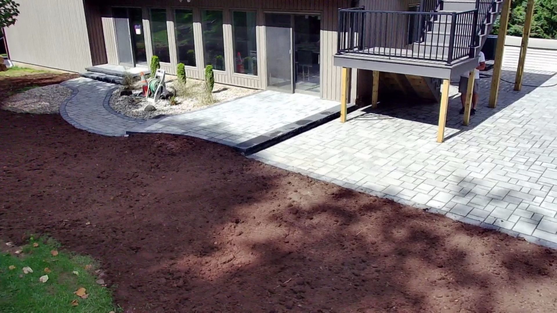 Modern paver patio with raised deck and black railing beside landscaped yard.