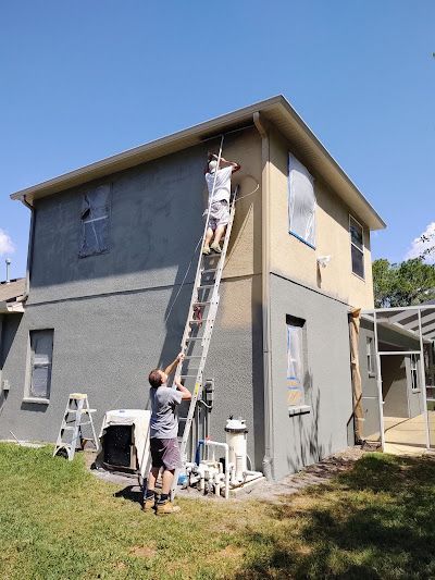 Two people painting a two-story house. One on a ladder applying paint, the other holding the ladder. Sunny day.