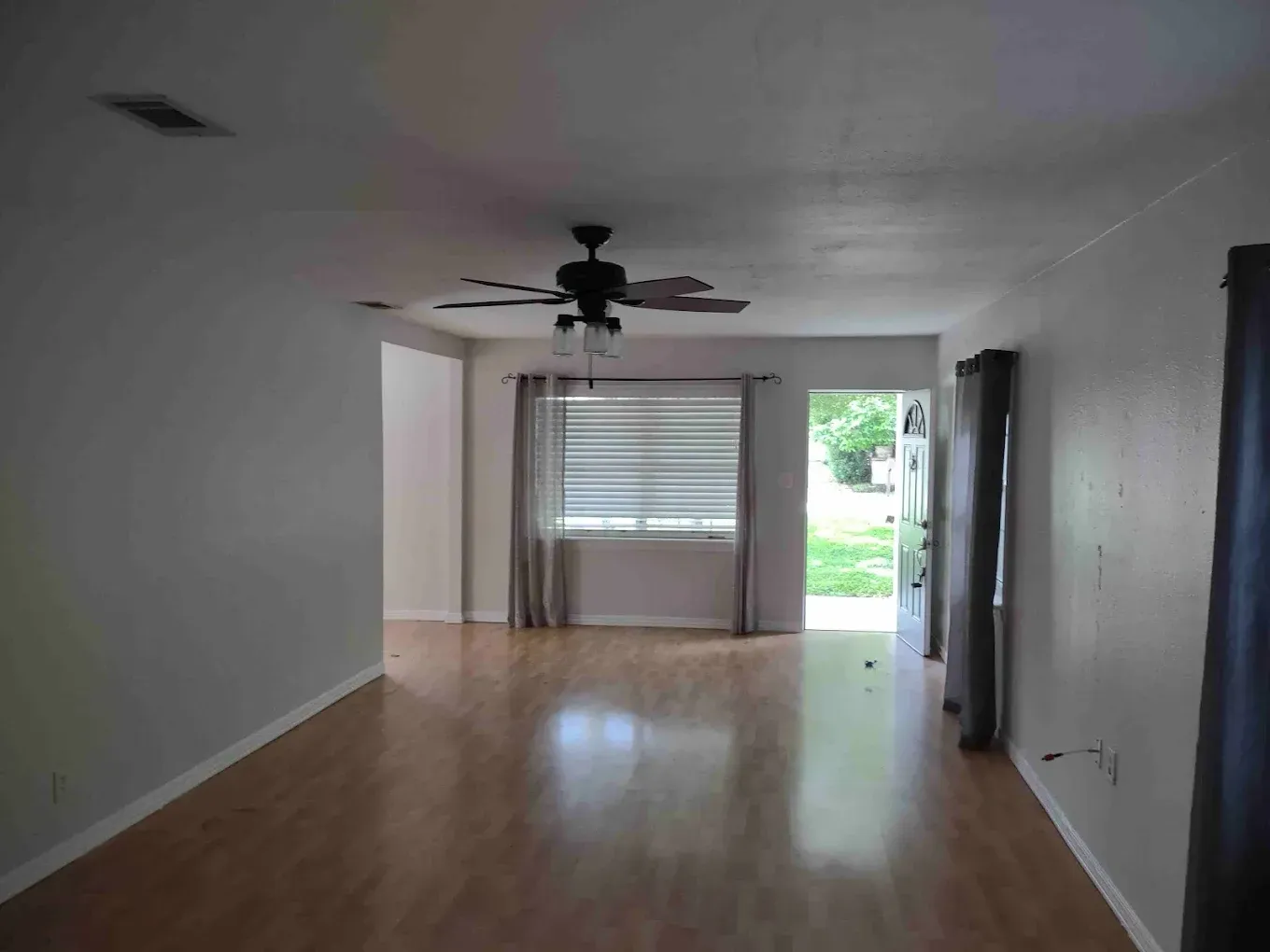 Interior view of a living room with laminate flooring, white walls, and a window with blinds.
