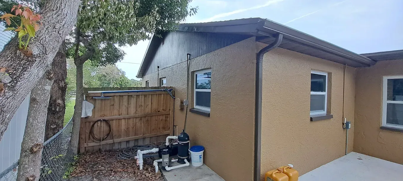 Exterior view of a stucco house with a wooden fence and pool equipment. Brown and beige tones.