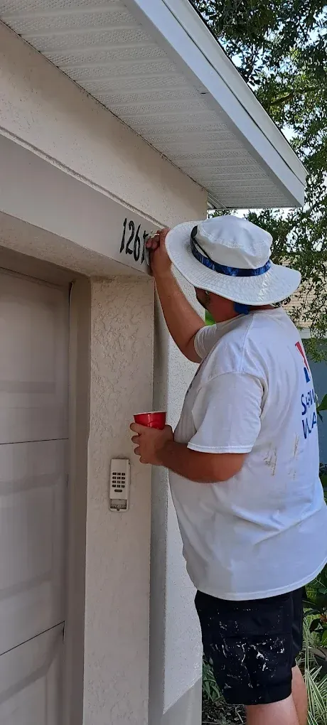 Person painting numbers on a building near the door while holding a cup, wearing a white hat, shorts, and a white t-shirt.