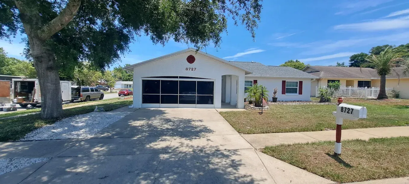 White suburban house with a dark garage door, a mailbox, and a tree in the foreground.