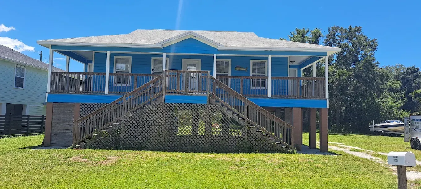 Blue house on stilts with wooden stairs, porch, and a grassy lawn.