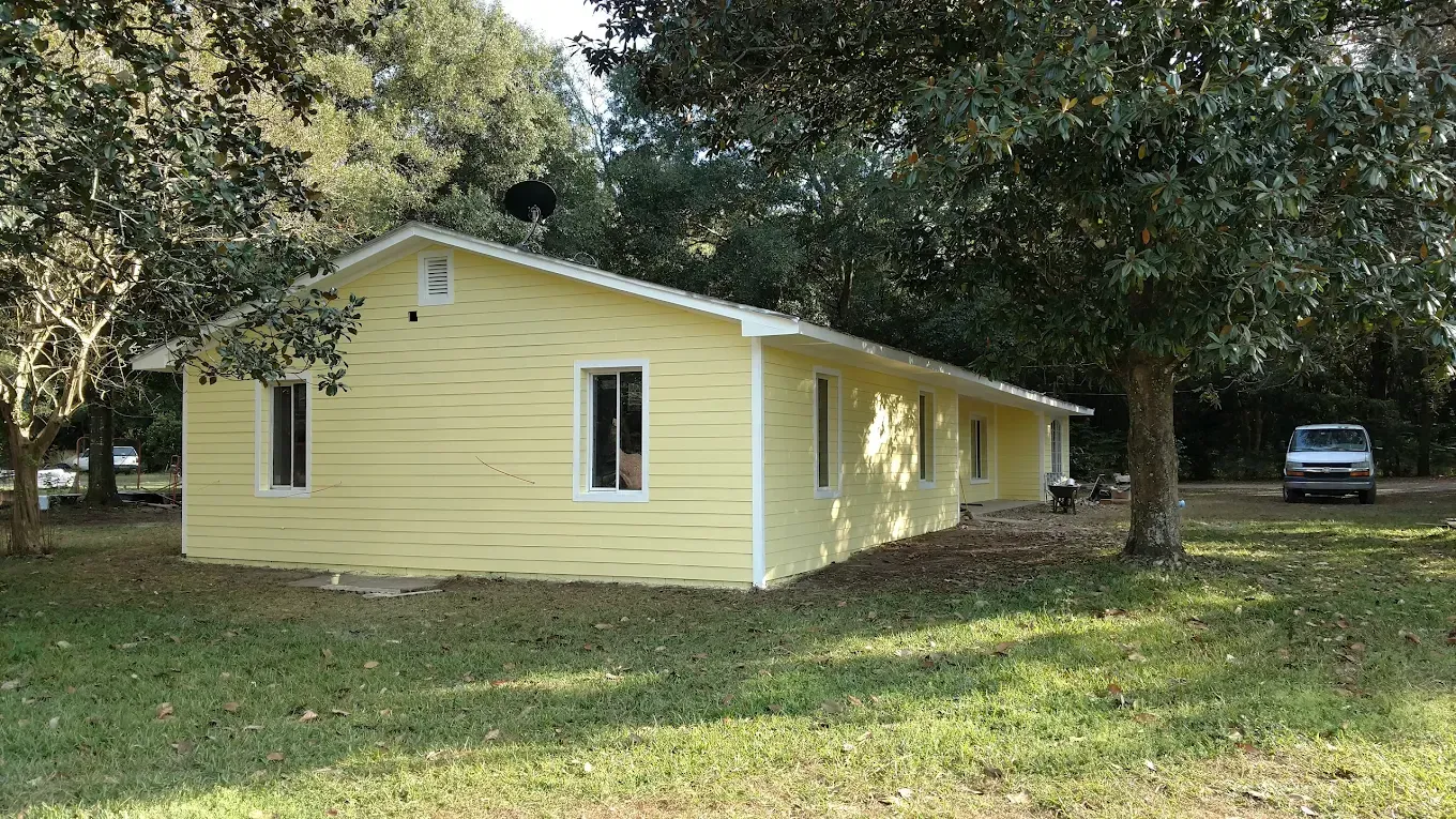Yellow house with white trim, set in a grassy yard, with a van parked nearby, and trees in the background.