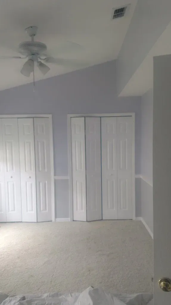 Bedroom with white bi-fold closet doors, light blue walls, and beige carpet. Ceiling fan visible.