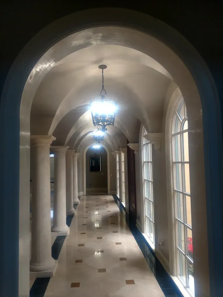 Hallway with arched ceiling, columns, and windows, lit by chandeliers. Marble floor with dark accents.