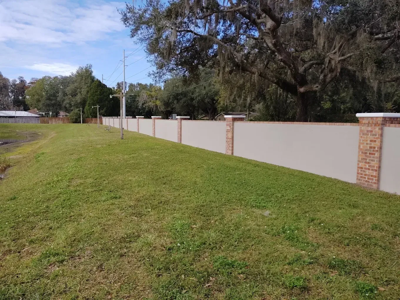 A long, light-colored wall with brick columns alongside green grass under a blue sky with trees.