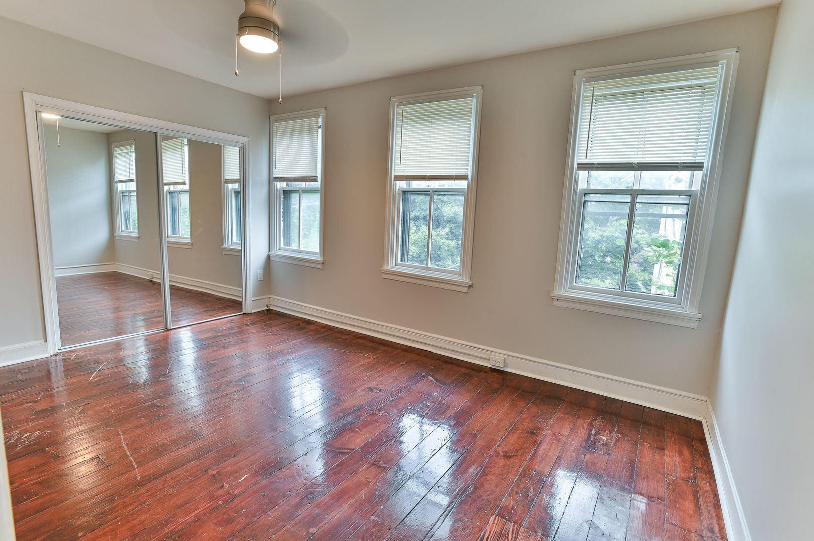 Empty room with hardwood floors, three windows with blinds, and mirrored closet doors.