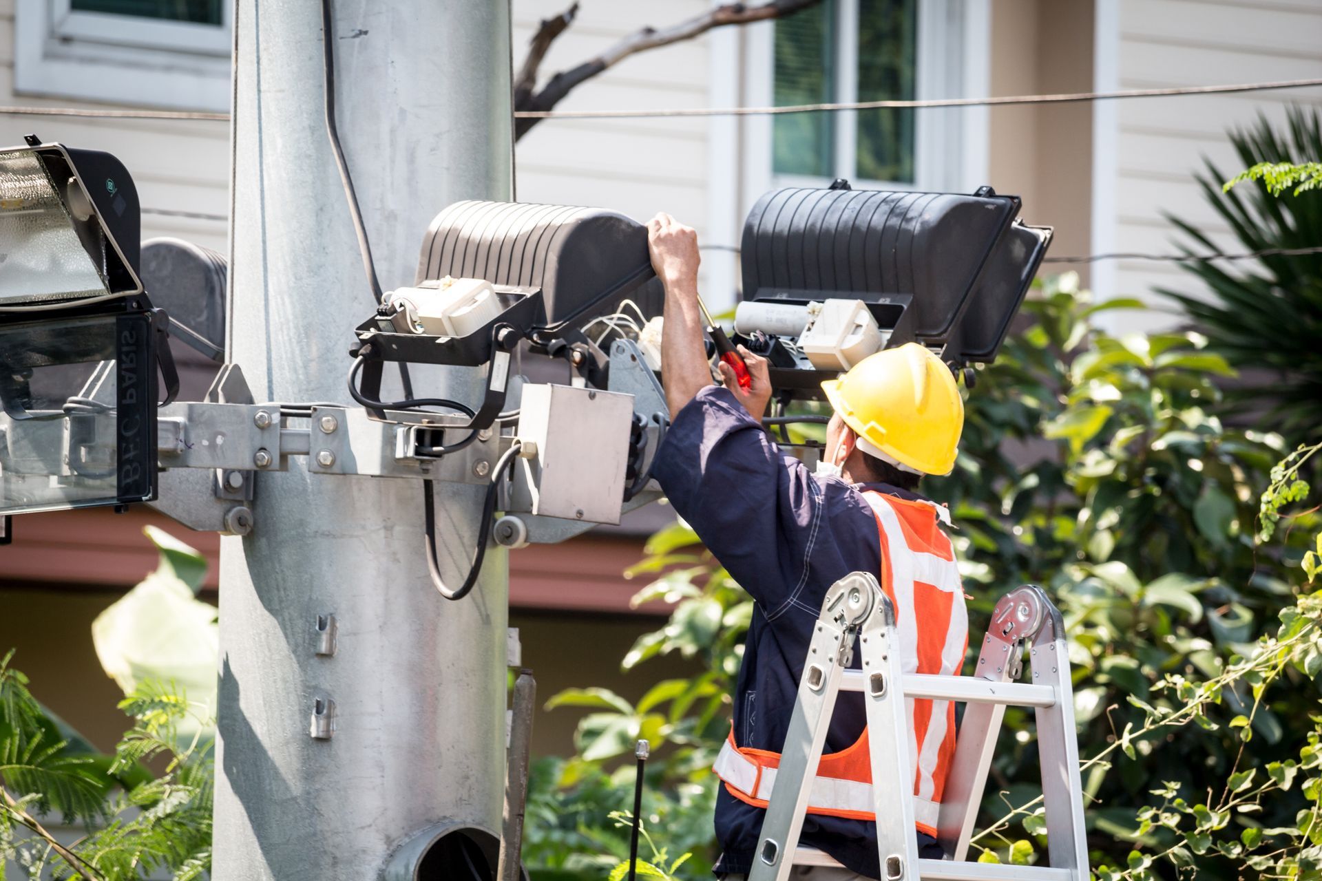 Lineman in safety gear working on a utility pole, using tools, with a house in the background.